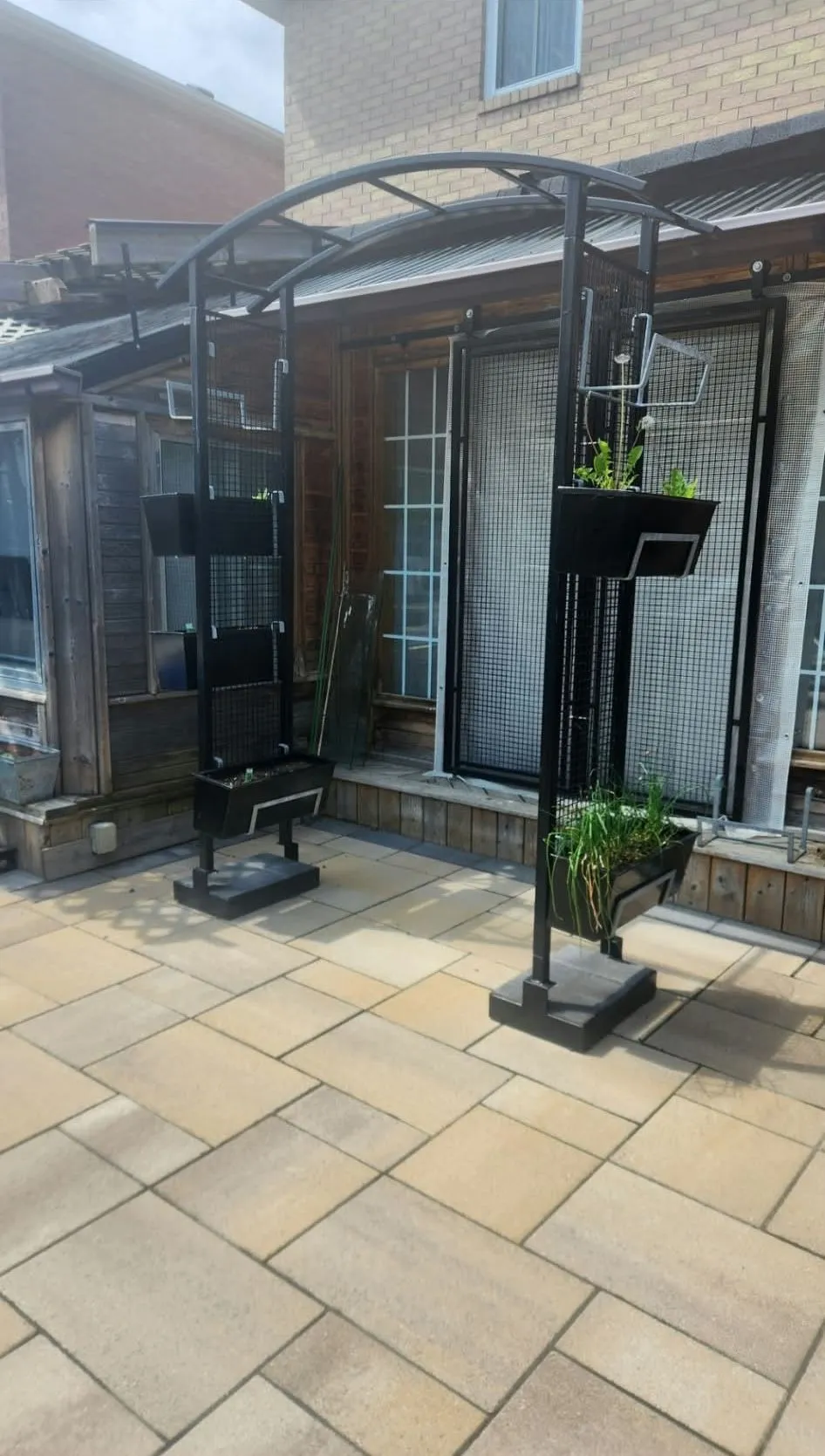 Two tall black metal posts joined by an arched top, holding hanging planter boxes with greenery, on a stone patio