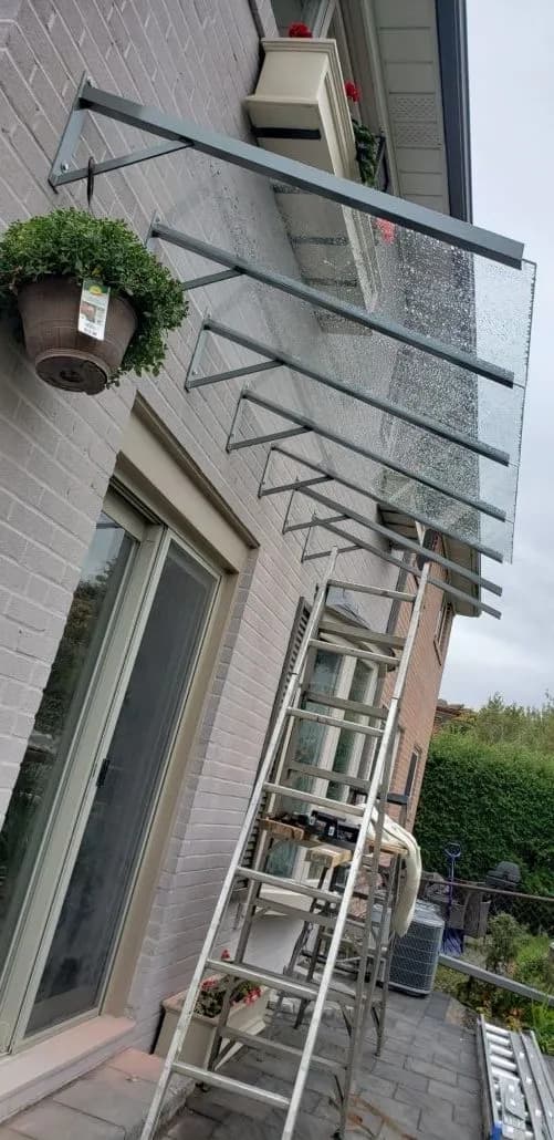 Glass canopy panels on steel arms above a patio door, with a ladder against the wall during installation