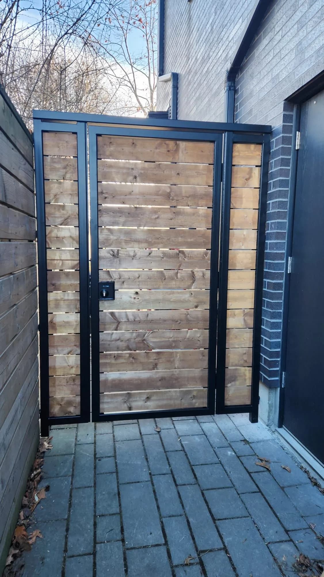 Black steel-framed walk-through gate with horizontal natural wood boards closing a narrow side passage