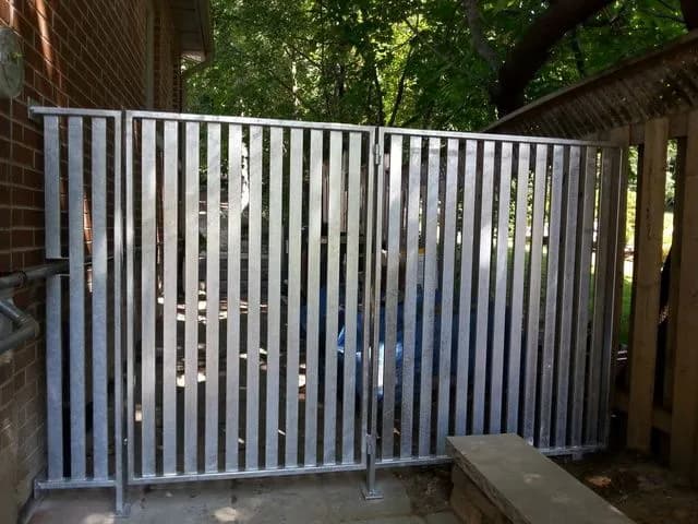 Galvanized steel gate and fence with tall vertical slats closing off a backyard corner beside a brick house