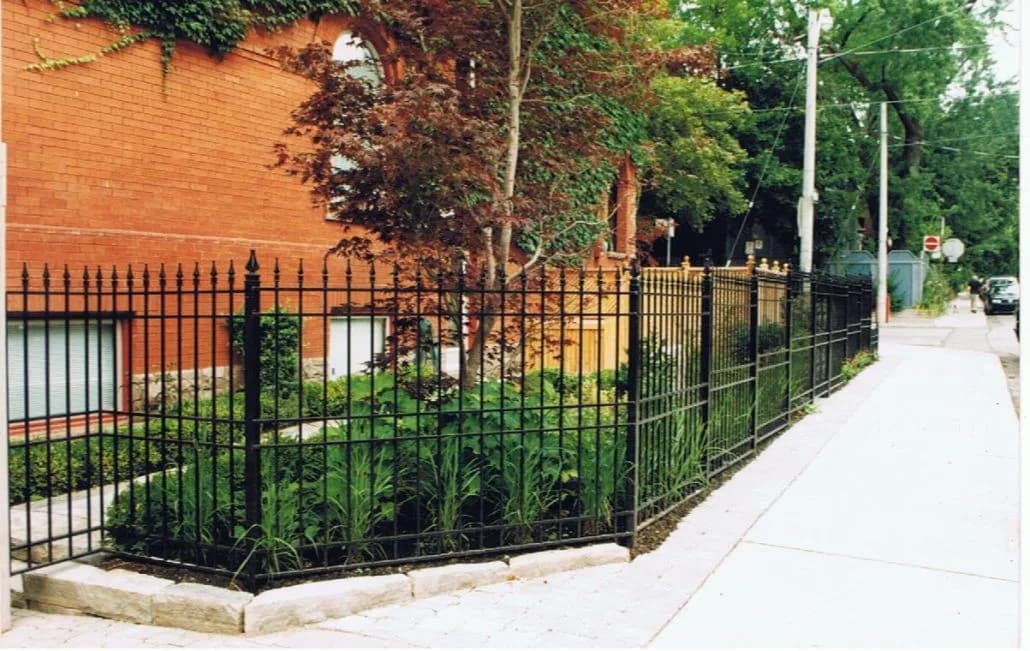 Black steel picket fence with finials enclosing a planted front garden along a sidewalk beside a brick building