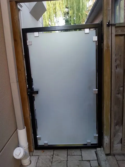 Walk-through gate made of a frosted glass panel in a black steel frame, set between a house wall and a wood fence