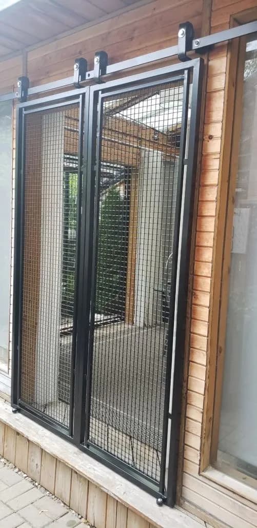 Pair of black steel sliding doors with wire mesh infill hung on an overhead track across a cedar-clad enclosure
