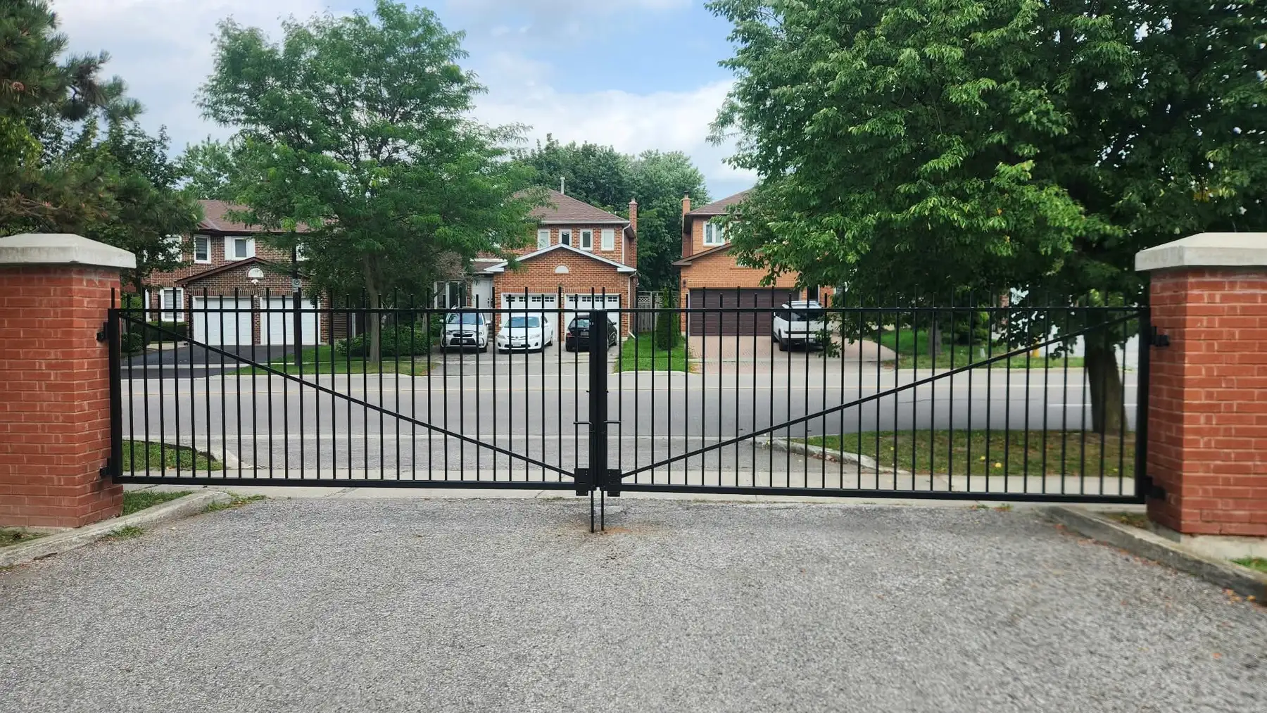 Wide black metal double driveway gate with vertical pickets and diagonal braces hung between two red brick pillars