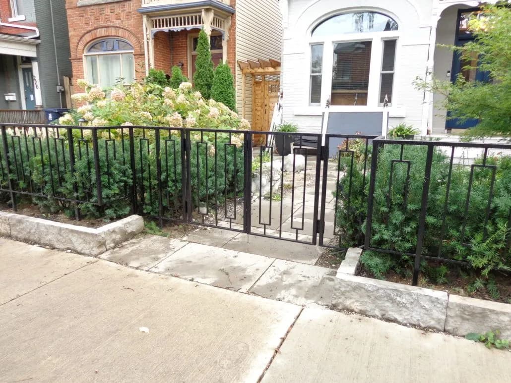 Black iron picket fence with a walk-through gate running along a hedged front garden beside a sidewalk