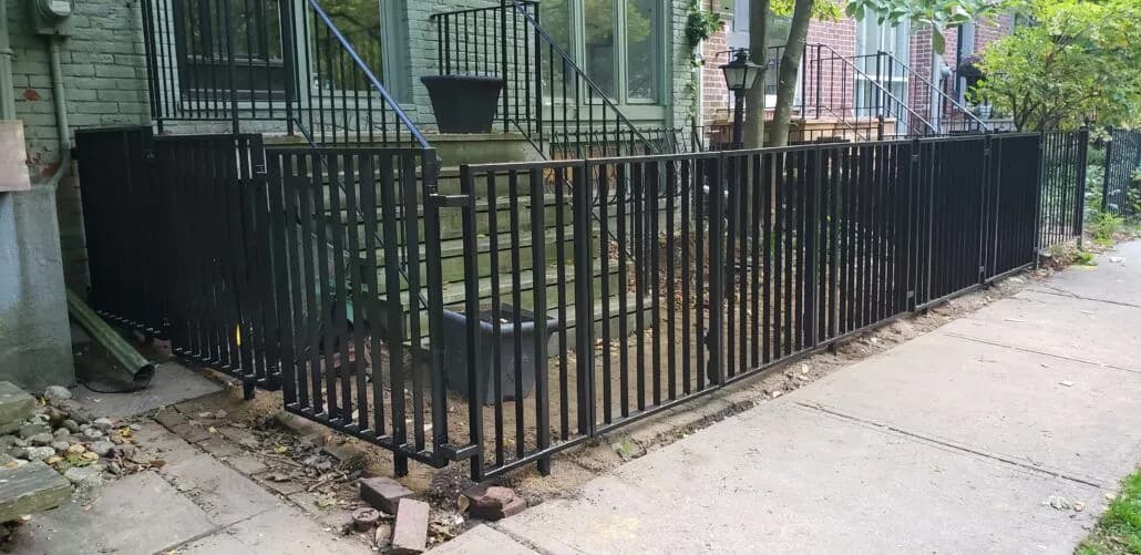 Black picket fence with finial tops enclosing a front yard, running along a city sidewalk
