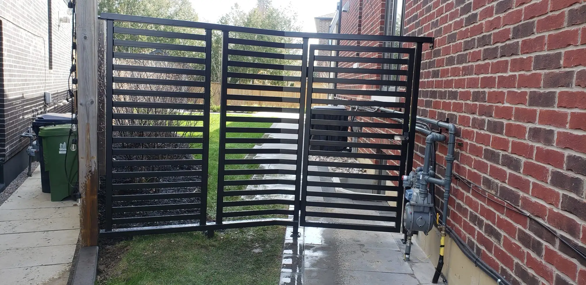 Two black horizontal slat gates across a side passage between a brick wall and a fence
