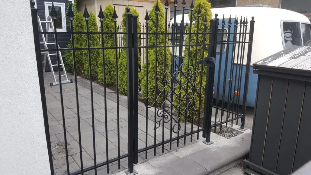 Black spear-top iron gate and matching fence panel beside a paved walkway lined with cedars