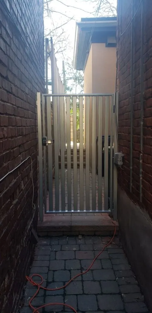 Black steel picket gate with a latch across a narrow brick side passage over interlock paving