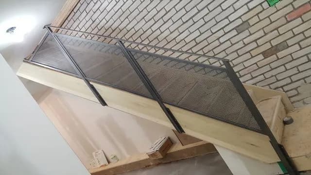 Black perforated metal guard panels along a plywood staircase during construction beside a brick wall