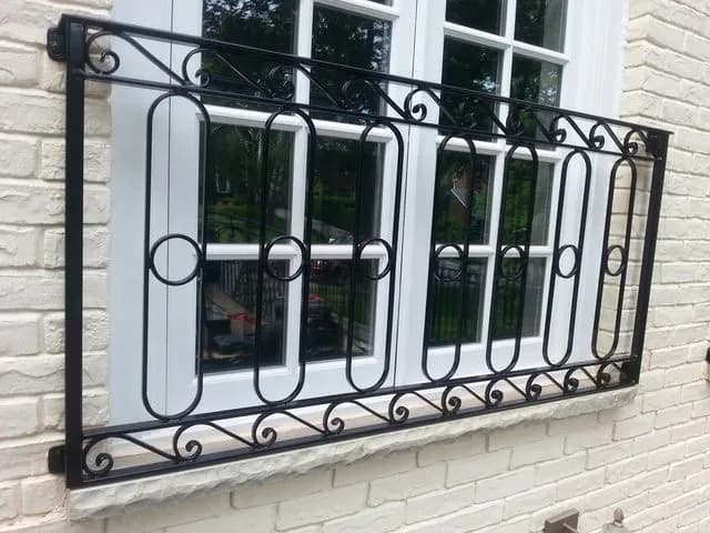 Black wrought iron Juliet railing with oval loops and scrolls fixed across a tall white-framed window on light brick