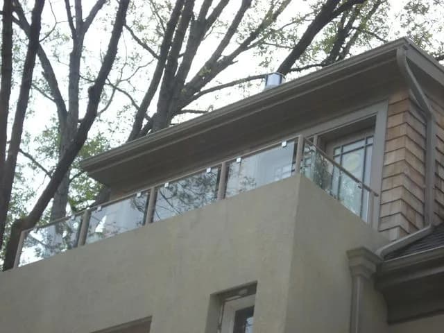 Frameless glass railing across an upper-floor balcony opening on a stucco house among trees