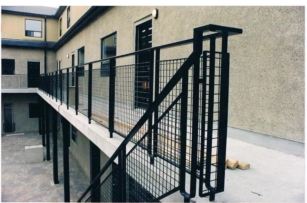 Black steel exterior stair and walkway guards with welded wire mesh panels along the upper level of a stucco courtyard