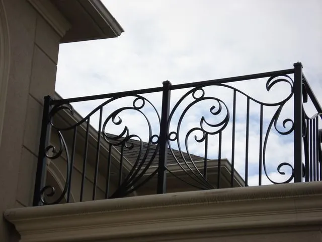 Black iron railing with large open scrolls between vertical bars along the edge of a stone balcony against the sky