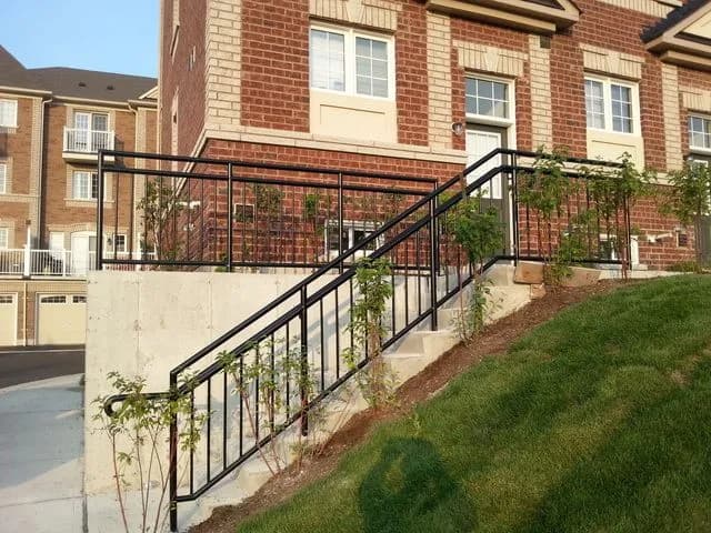 Black steel railings along a concrete exterior staircase and retaining wall in front of a row of brick townhouses