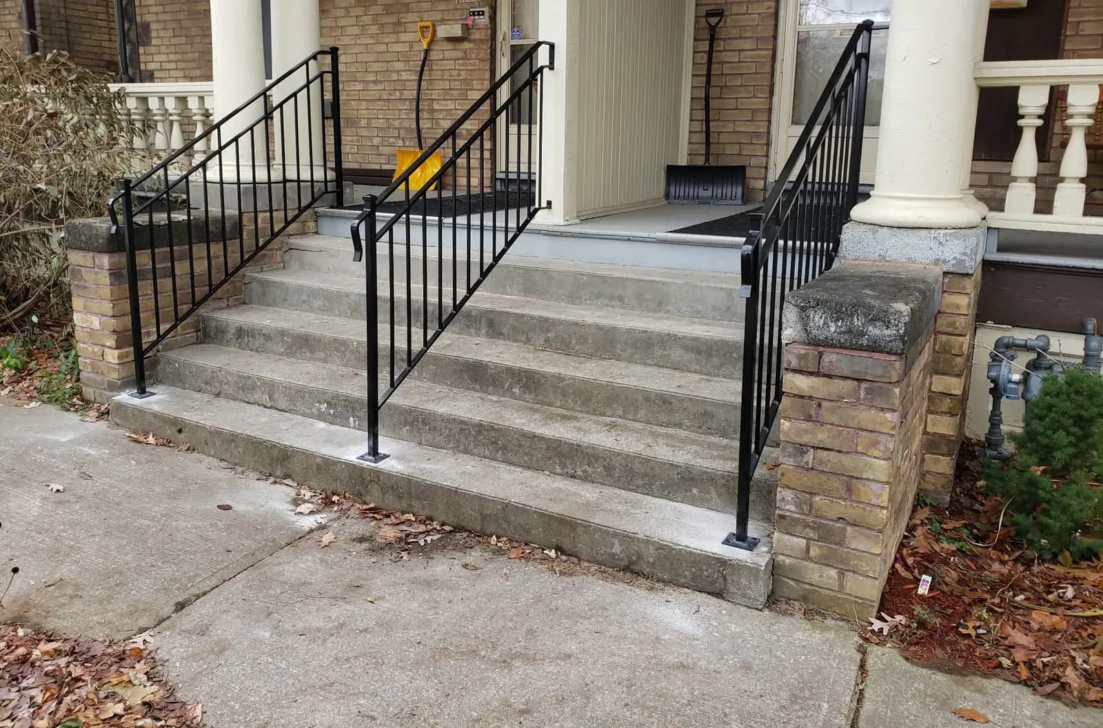 Three black metal picket railings on wide concrete front steps leading up to a porch with white columns and brick piers