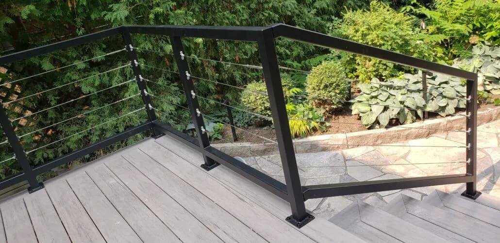 Horizontal cable railing between black posts with a wide top rail around a composite deck beside a garden