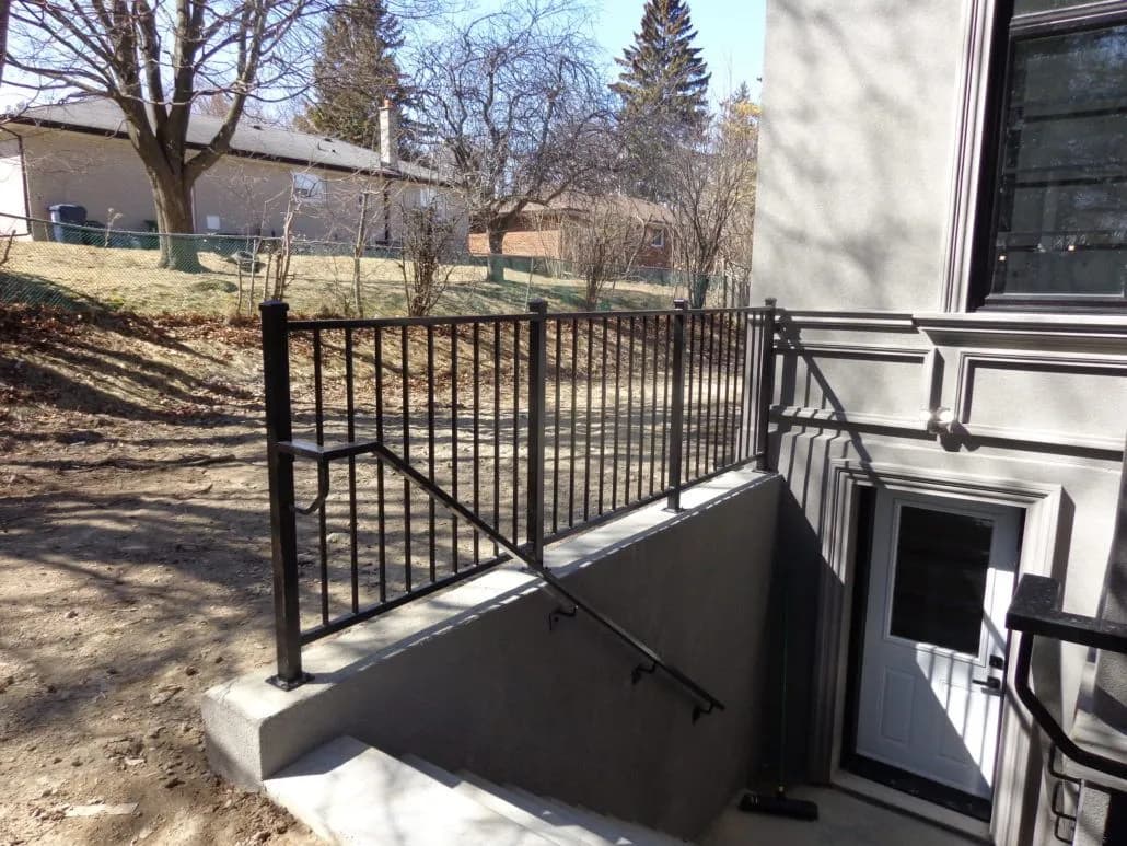 Black metal railing with vertical pickets around an outdoor basement stairwell beside a house