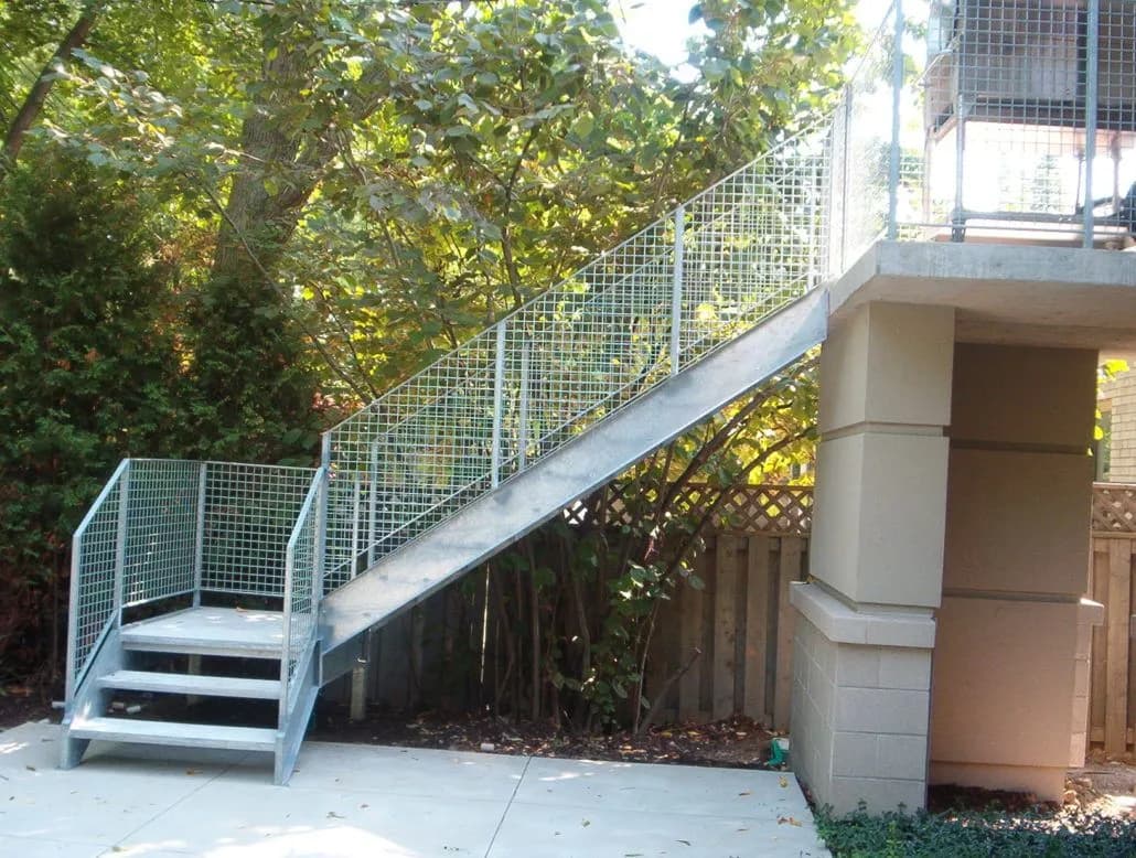 Galvanized steel exterior stair with mesh guards and a landing climbing along a stucco wall under trees