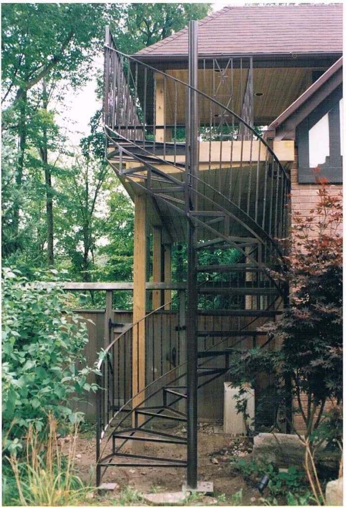 Black spiral staircase climbing to an upper-floor deck at the back of a house among trees
